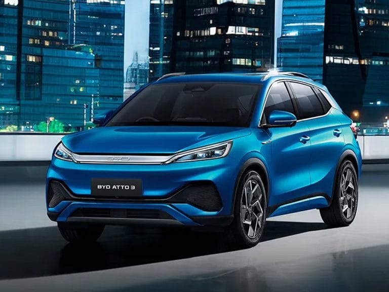 A blue BYD Atto 3 electric SUV is parked in front of a modern cityscape at night, with illuminated skyscrapers in the background.